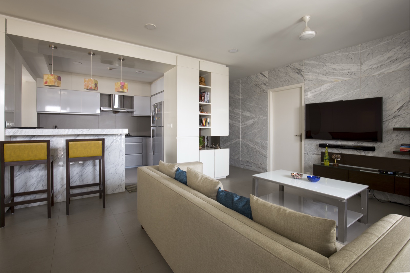 Living room with a marble television feature wall, beige sectional sofa and open modular kitchen by WOODWORKS Interior, Bengaluru