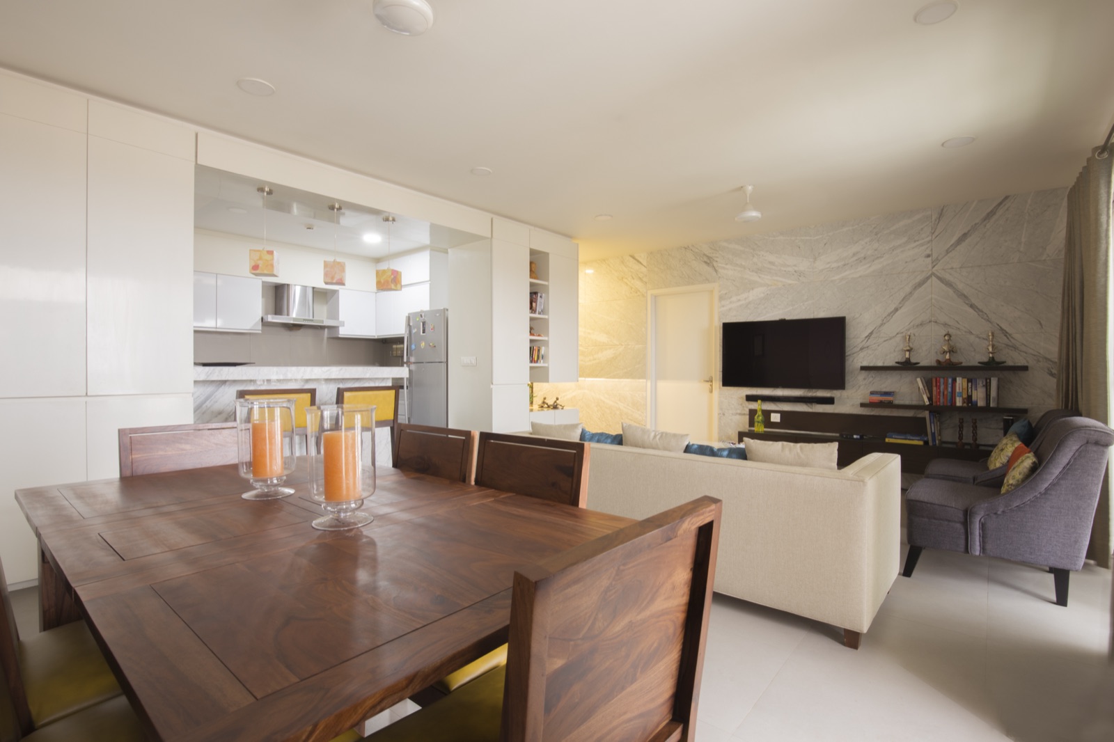 Open-plan living and dining space with a stone feature wall, solid-wood dining table and open modular kitchen by WOODWORKS Interior, Bengaluru
