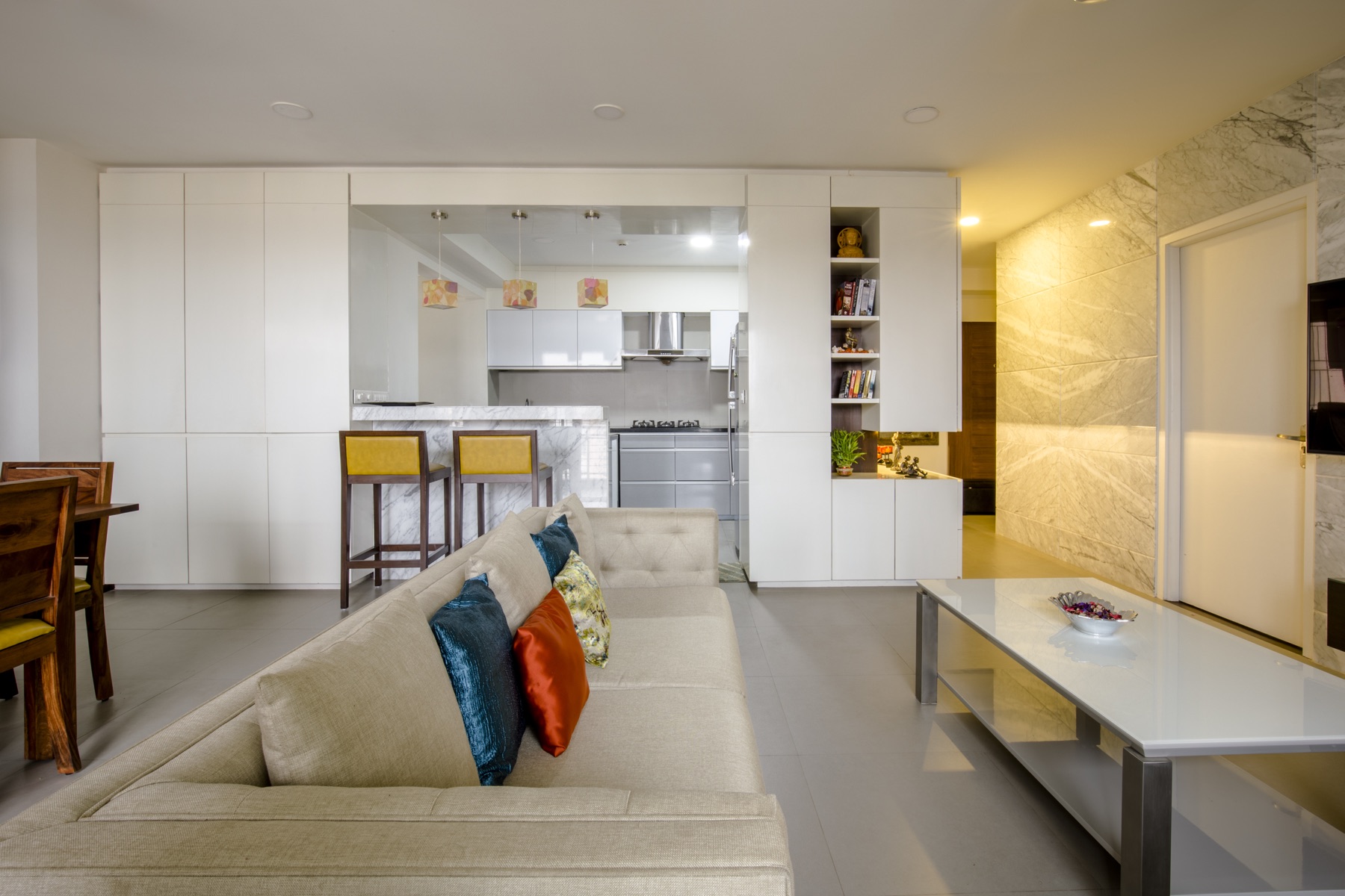 Open-plan modular kitchen with a veined-marble island, yellow bar stools and a bright living area by WOODWORKS Interior, Bengaluru