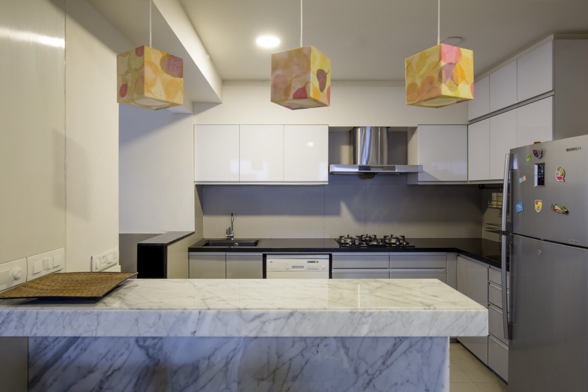 Modular kitchen with glossy white cabinets, a veined marble breakfast island and a trio of patterned pendant lights by WOODWORKS Interior, Bengaluru