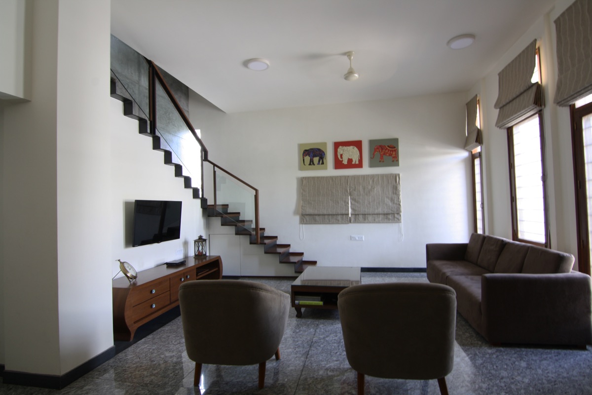 Double-height living room with a floating teak-and-glass staircase and plush lounge seating under tall daylit windows by WOODWORKS Interior, Bengaluru
