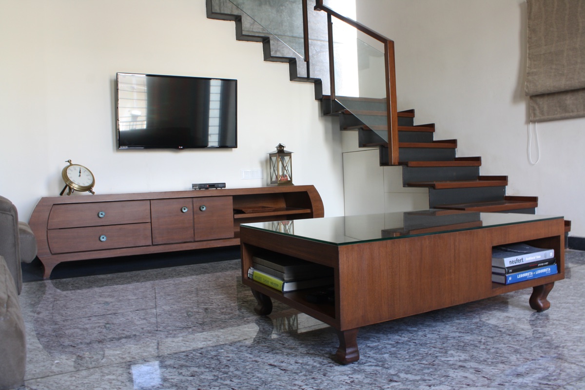 Bespoke walnut media console and glass-topped coffee table beside a cantilevered wood staircase by WOODWORKS Interior, Bengaluru